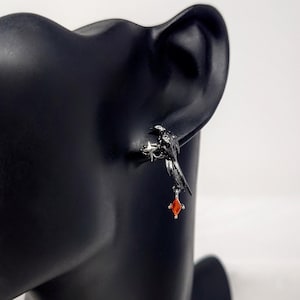 May include: A black raven earring with a silver-colored star-like base and a dangling red gemstone. The bird is perched on a silver-colored base. The earring is displayed on a black mannequin head.