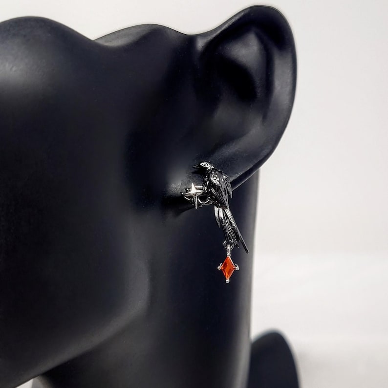 May include: A black raven earring with a silver-colored star-like base and a dangling red gemstone. The bird is perched on a silver-colored base. The earring is displayed on a black mannequin head.