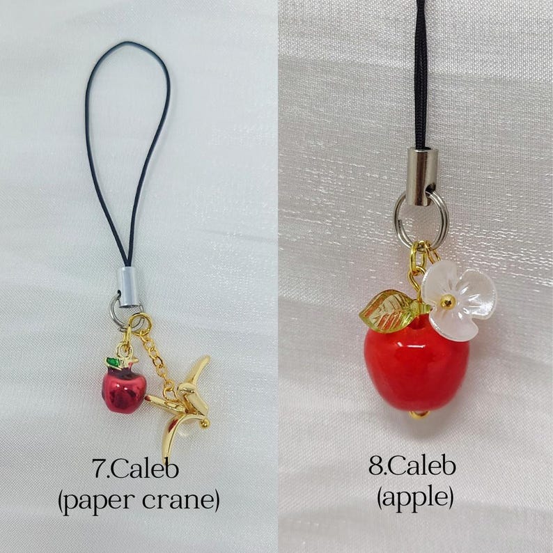 May include: Two phone charms with black cords. The first, labeled 7.Caleb, features a small red apple charm and a gold-toned metal paper crane. The second, labeled 8.Caleb, features a red apple charm with a green leaf and a white flower accent.
