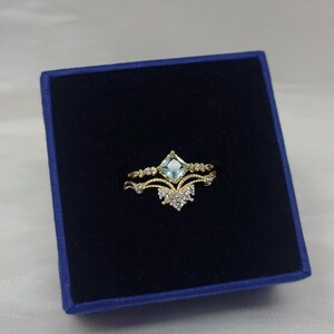 May include: A gold-toned ring with a light blue square gemstone at the center, set in a crown-like design. The ring features delicate detailing and is presented in a dark blue box, showcasing its elegant design.