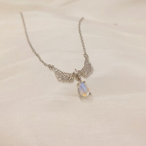 May include: A silver necklace with a teardrop-shaped opal pendant and angel wings adorned with clear cubic zirconia stones.