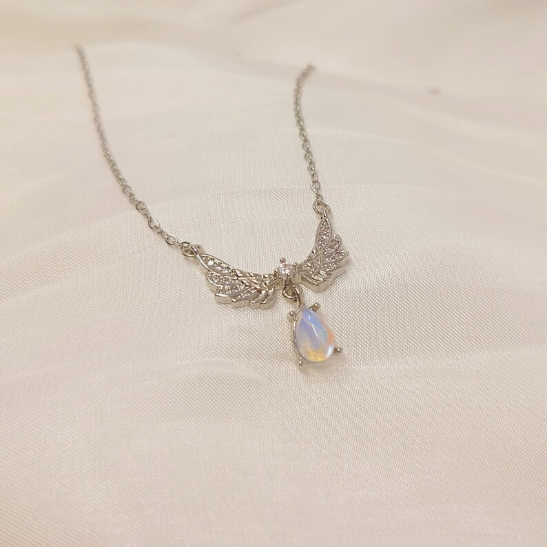May include: A silver necklace with a teardrop-shaped opal pendant and angel wings adorned with clear cubic zirconia stones.