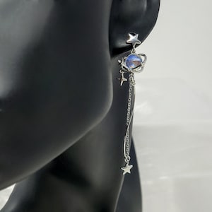 Puede incluir: Pendientes colgantes plateados con tem&aacute;tica celestial. Cada pendiente presenta un orbe similar a un planeta con un tono azul y morado, rodeado por un anillo y peque&ntilde;as estrellas. Una cadena con un dije de estrella cuelga debajo.
