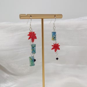 May include: Dangle earrings featuring red maple leaf charms and cylindrical beads with blue and green patterns. The earrings are suspended from silver-colored hooks and chains, with a small black bead at the bottom of one earring.