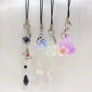 May include: Four phone charms with different designs. The first charm features a black rose with a silver butterfly and a black crystal. The second charm features a blue and white flower with a silver chain and white pearls. The third charm features a white and pink flower with a silver leaf. The fourth charm features a purple flower with a pink flower on top.