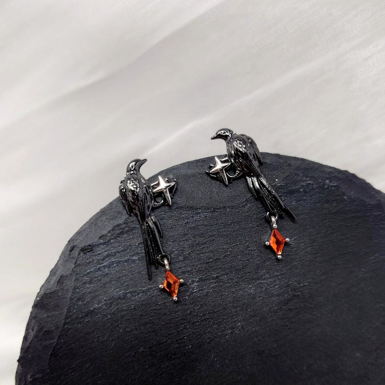 May include: A pair of black raven earrings. Each earring features a raven perched on a star, with a dangling orange gemstone. The earrings are set against a dark, textured surface.