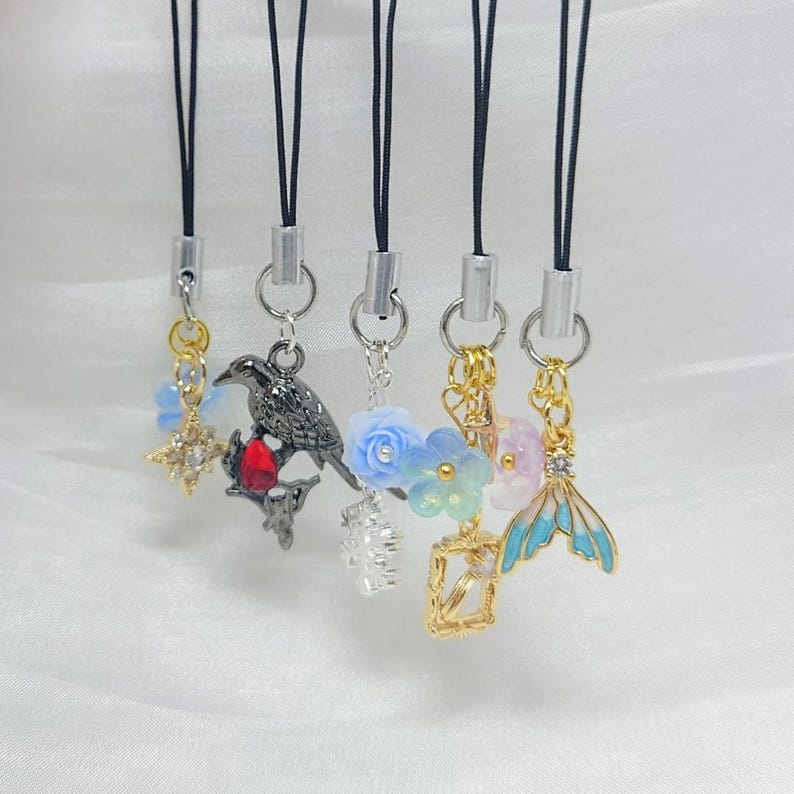 May include: Five phone charms hanging from black cords with silver-tone connectors. Charms include a gold star, a black bird with a red stone, a blue flower, a gold frame with a blue flower, and a blue and gold mermaid tail with a clear stone.