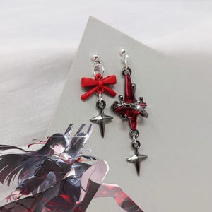 May include: A pair of dangle earrings featuring a red bow and a star-shaped charm. One earring has a red starburst design. The earrings are silver-toned and have a post closure. The earrings are on a white background.