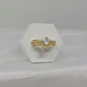 May include: A gold-colored ring set with an aquamarine-colored, square-cut gemstone and small white gemstones. The ring has a delicate, ornate design. The set includes a band and a crown-shaped ring.