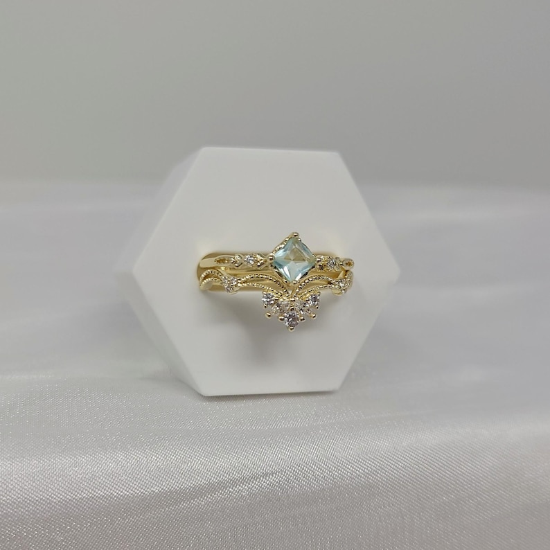 May include: A gold-colored ring set with an aquamarine-colored, square-cut gemstone and small white gemstones. The ring has a delicate, ornate design. The set includes a band and a crown-shaped ring.