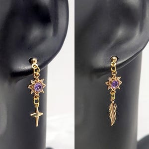 May include: Gold-toned earrings with a starburst design featuring a purple center stone and small clear accents. One earring has a dangling gold cross, the other a gold feather. The earrings are displayed on a black earring holder.
