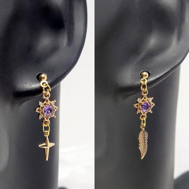 May include: Gold-toned earrings with a starburst design featuring a purple center stone and small clear accents. One earring has a dangling gold cross, the other a gold feather. The earrings are displayed on a black earring holder.