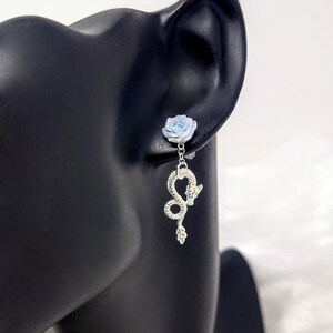 May include: A silver earring featuring a light blue rose stud and a dangling snake charm. The snake charm is detailed with a textured design and a small leaf at the bottom. The earring is displayed on a black mannequin head.