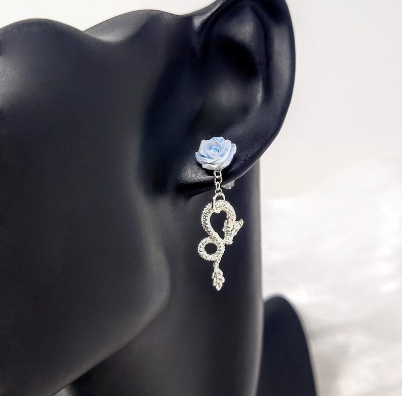 May include: A silver earring featuring a light blue rose stud and a dangling snake charm. The snake charm is detailed with a textured design and a small leaf at the bottom. The earring is displayed on a black mannequin head.