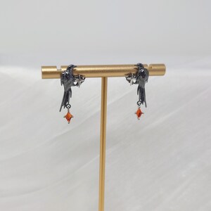 May include: A pair of black raven earrings with dangling orange gemstone accents. The ravens are perched on a gold-colored bar, and the earrings are displayed against a soft, white background. The earrings are a unique and stylish accessory.