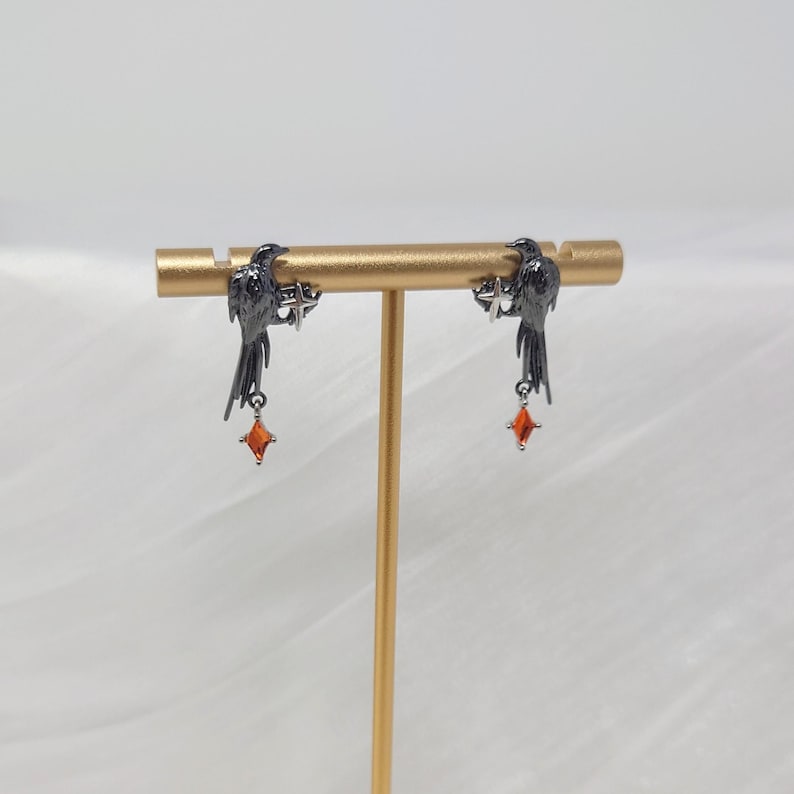 May include: A pair of black raven earrings with dangling orange gemstone accents. The ravens are perched on a gold-colored bar, and the earrings are displayed against a soft, white background. The earrings are a unique and stylish accessory.