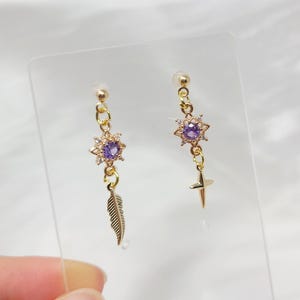 May include: A pair of gold-colored earrings. One earring features a purple gemstone in a starburst design, with a dangling feather charm. The other earring has a similar starburst design with a purple gemstone and a dangling gold-colored star charm.