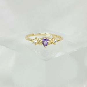 May include: A gold-colored ring with a heart-shaped, purple gemstone at the center. The ring is flanked by two smaller, clear gemstones. The band is adjustable and features delicate decorative details.