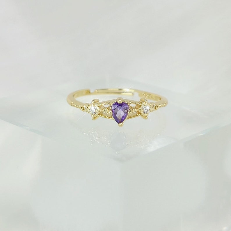 May include: A gold-colored ring with a heart-shaped, purple gemstone at the center. The ring is flanked by two smaller, clear gemstones. The band is adjustable and features delicate decorative details.