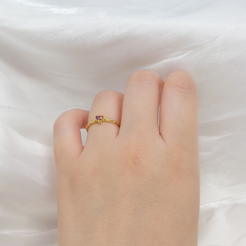 K&ouml;nnte beinhalten: Ein goldener Ring mit einem kleinen, roten herzf&ouml;rmigen Edelstein. Der Ring hat eine strukturierte Band.
