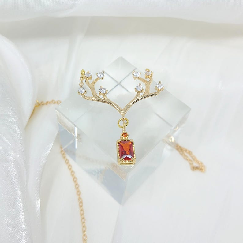May include: A gold-toned pendant necklace featuring a stylized antler design with clear crystal accents. The pendant includes a rectangular, red gemstone. The necklace is displayed on a clear, geometric block.