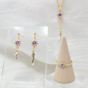 May include: A gold-toned jewelry set featuring a necklace, earrings, and a ring. The earrings have a cross and feather charm. The ring and necklace have a purple gemstone. The jewelry is displayed on a neutral background.