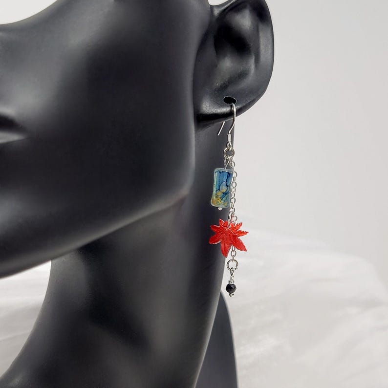 May include: Silver-tone dangle earrings featuring a rectangular blue and yellow charm, a bright red maple leaf, and a small black bead. The earrings are designed with a chain and hook closure, showcasing a Japanese-inspired aesthetic.