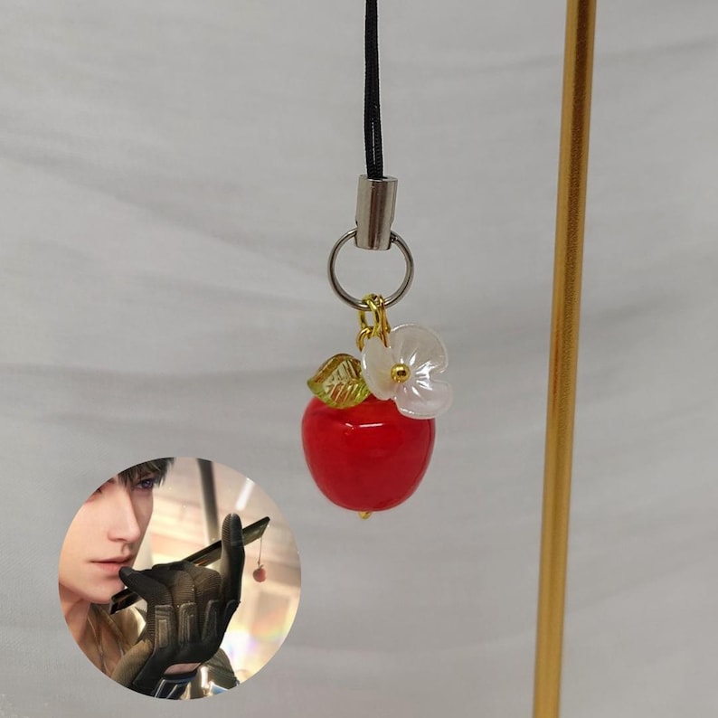 May include: A phone charm featuring a glossy red apple pendant, a small translucent green leaf, and a white flower-shaped bead. The charm hangs from a silver-toned metal ring attached to a black cord. An inset shows a character holding a similar charm.