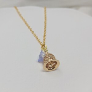 May include: A gold-colored necklace with a bell-shaped pendant. The pendant has intricate cut-out designs and is adorned with a small blue flower and a white pearl. The chain is delicate, and the overall design is elegant.