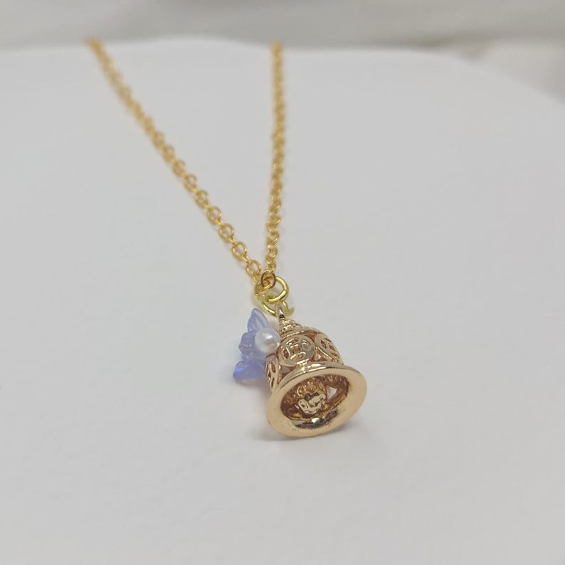 May include: A gold-colored necklace with a bell-shaped pendant. The pendant has intricate cut-out designs and is adorned with a small blue flower and a white pearl. The chain is delicate, and the overall design is elegant.