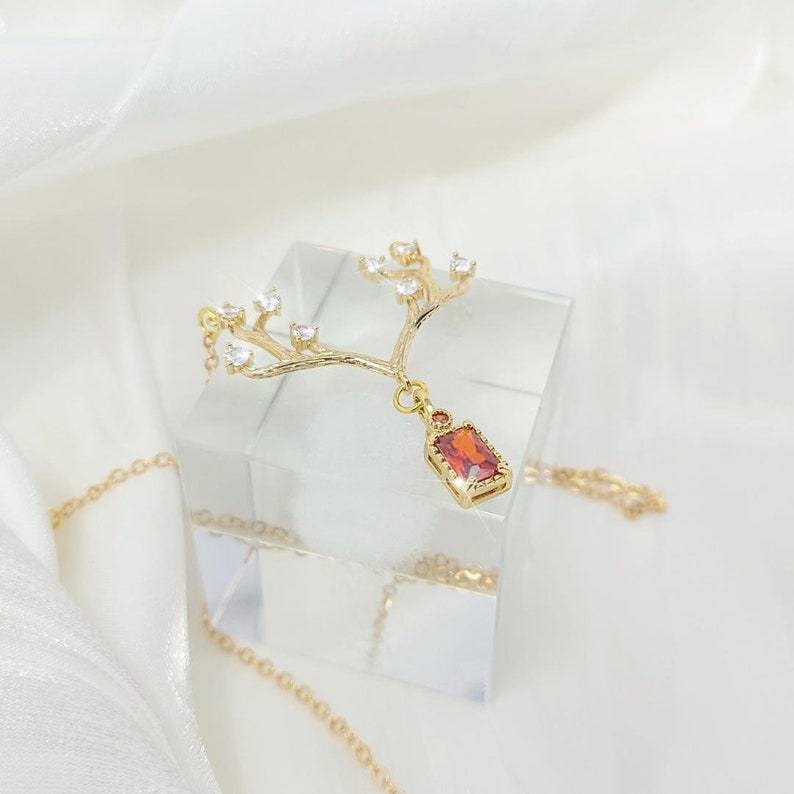 May include: A gold-colored necklace featuring a tree branch design with clear crystals and a rectangular, reddish-orange gemstone pendant. The necklace is displayed on a clear, square block against a white background.