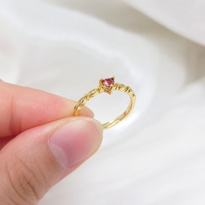 K&ouml;nnte beinhalten: Ein goldener Ring mit einem kleinen roten herzf&ouml;rmigen Edelstein und einem zarten Band aus winzigen klaren Steinen.