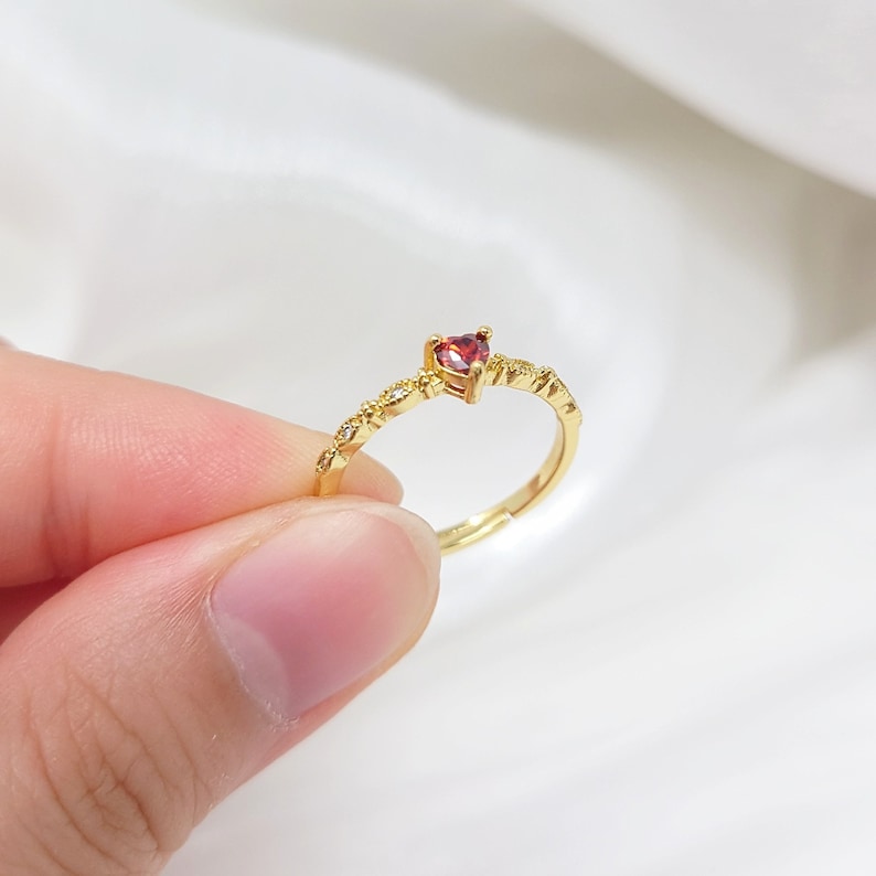 K&ouml;nnte beinhalten: Ein goldener Ring mit einem kleinen roten herzf&ouml;rmigen Edelstein und einem zarten Band aus winzigen klaren Steinen.