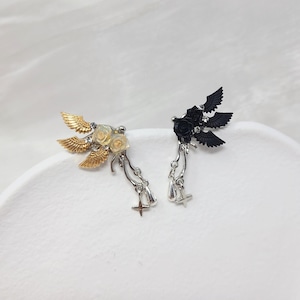 May include: A pair of decorative ear cuffs. One cuff features gold-colored wings and cream-colored roses, while the other has black wings and roses. Both have dangling silver-colored charms with crosses.
