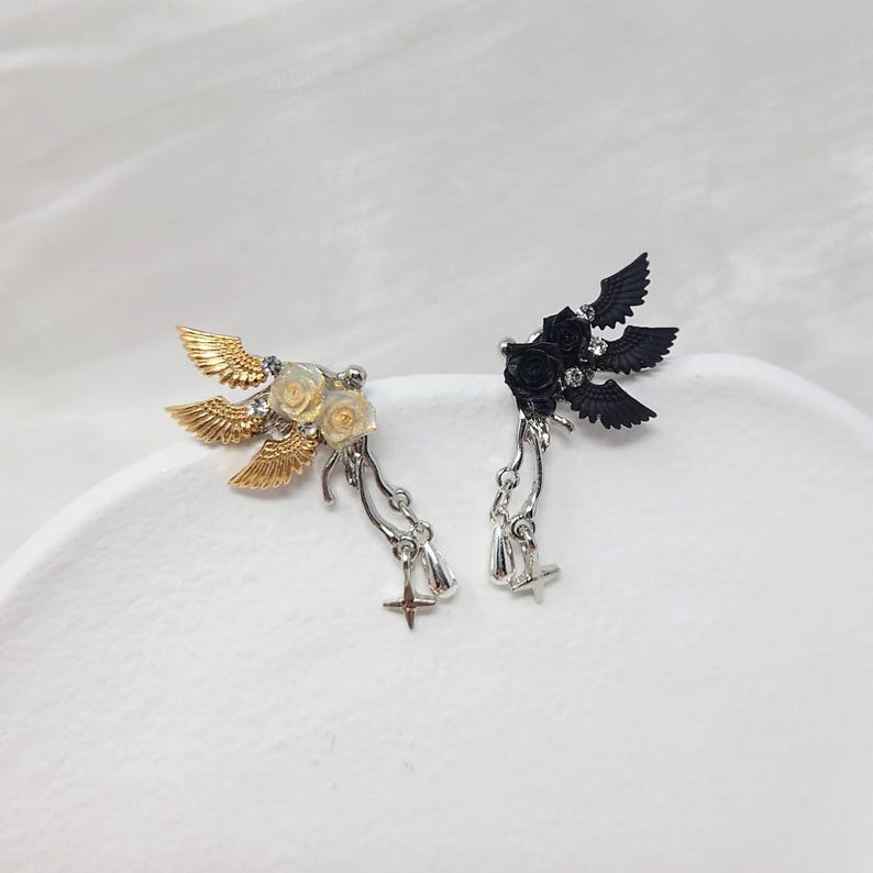 May include: A pair of decorative ear cuffs. One cuff features gold-colored wings and cream-colored roses, while the other has black wings and roses. Both have dangling silver-colored charms with crosses.