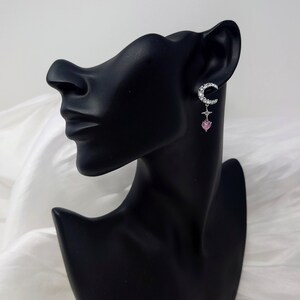 May include: Silver crescent moon earrings with a pink heart charm dangling from the bottom. The earrings are set with clear rhinestones.