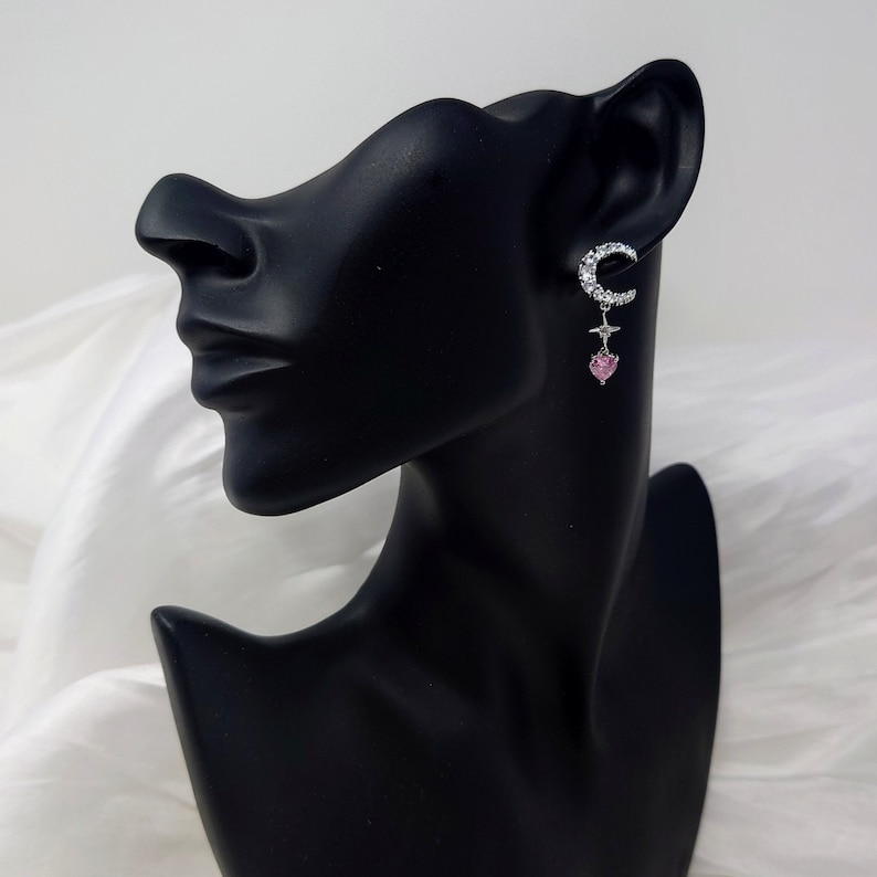 May include: Silver crescent moon earrings with a pink heart charm dangling from the bottom. The earrings are set with clear rhinestones.