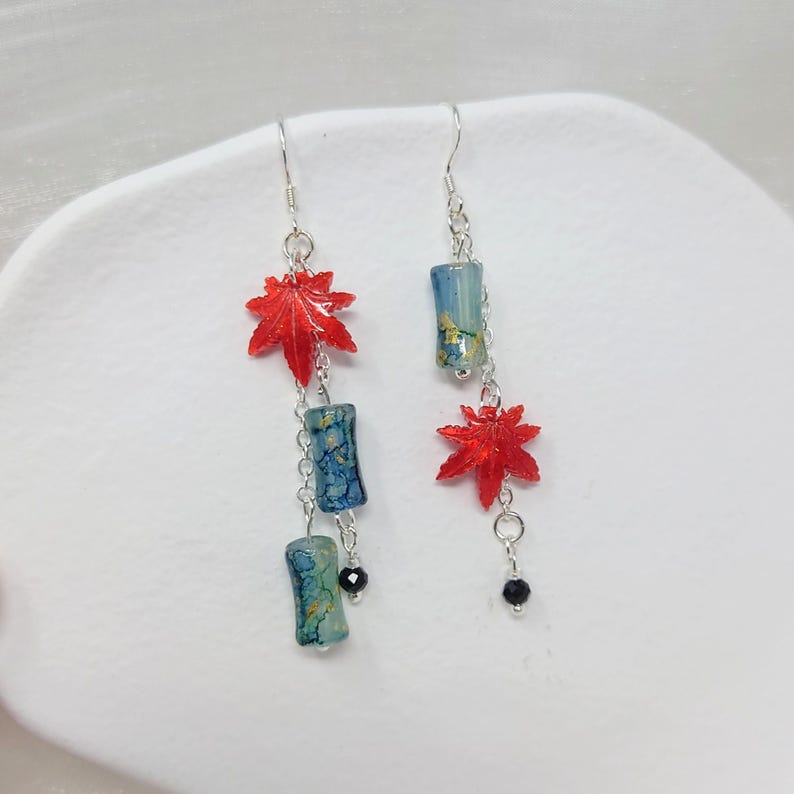 May include: Dangling earrings featuring red maple leaf charms, blue and gold cylindrical beads, and black accents. The earrings are suspended from silver hooks and chains, creating a delicate and eye-catching design. These earrings are a unique accessory.