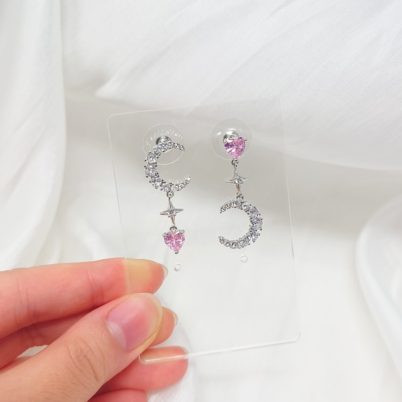 May include: A pair of silver earrings with a crescent moon, star, and pink heart charm. The earrings are displayed on a clear plastic backing.