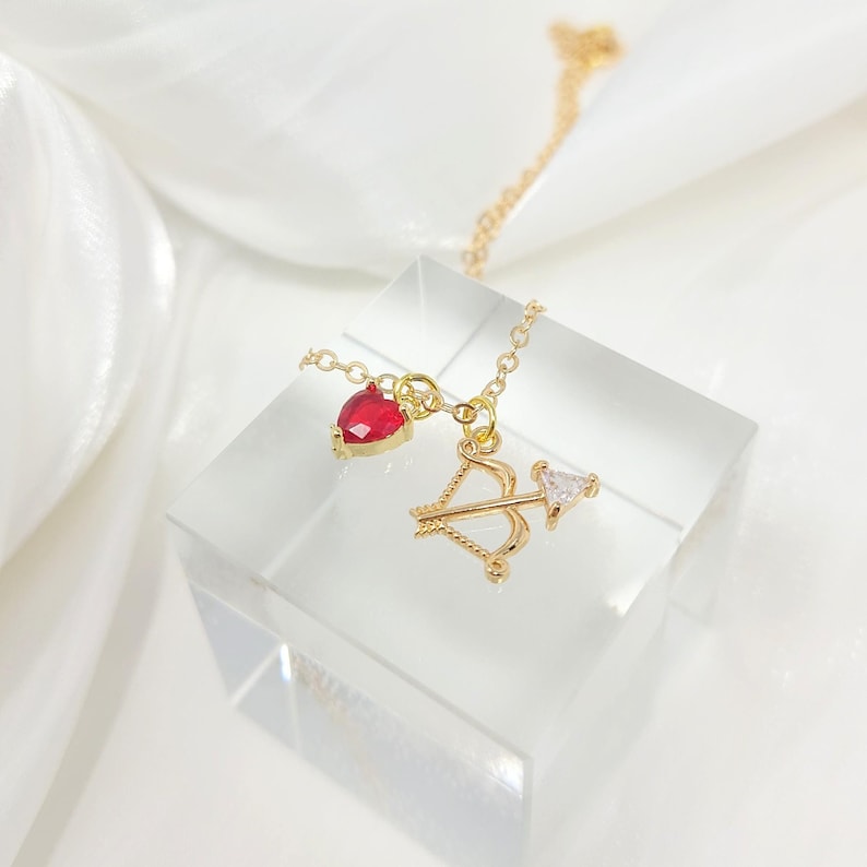 K&ouml;nnte beinhalten: Eine goldene Halskette mit einem roten Herz-Anh&auml;nger und einem Bogen-und-Pfeil-Anh&auml;nger. Die Halskette liegt auf einem wei&szlig;en Hintergrund.