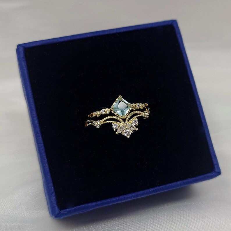 May include: A gold-colored ring set with a light blue square gemstone and small white accent stones. The ring is in a dark blue box, suggesting a jewelry presentation.