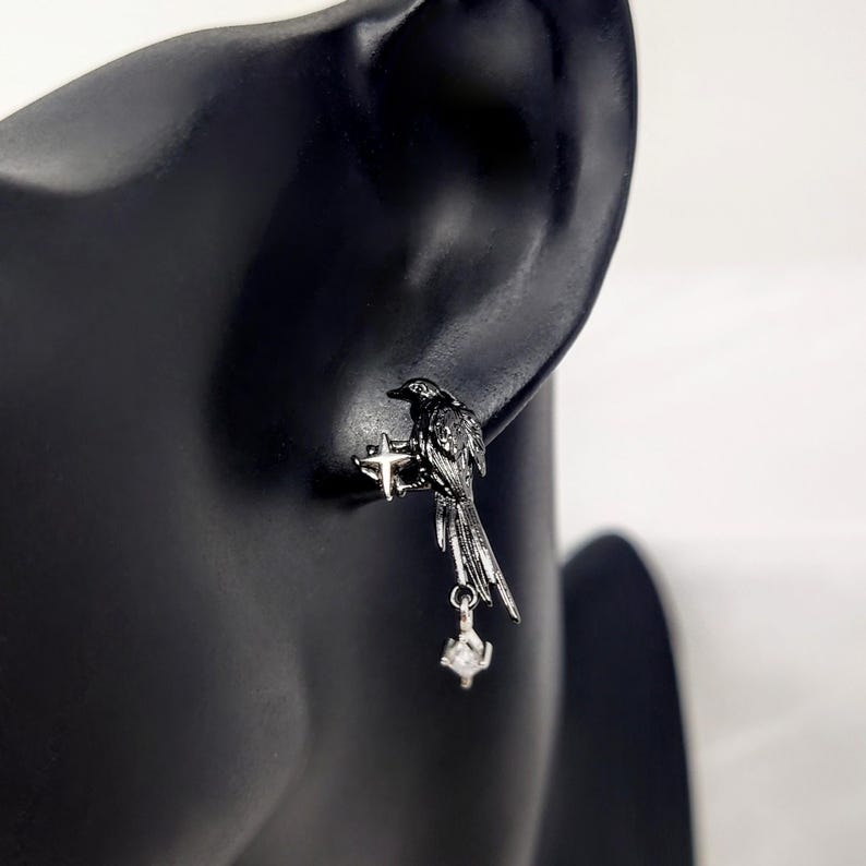May include: A pair of black raven earrings. Each earring features a detailed raven perched on a cross-shaped design, with a dangling, clear, cubic zirconia stone. The earrings are made of a dark metal and are displayed on a black mannequin.