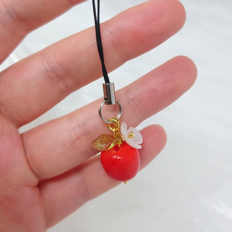 May include: A small charm featuring a bright red apple, a metallic gold leaf, and a white flower with a gold center. The charm is attached to a silver-toned jump ring and a black cord loop, designed for attachment to mobile devices or accessories.