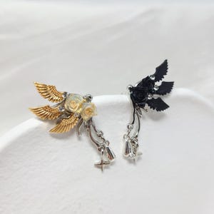 May include: Two decorative ear cuffs. One is gold-toned with gold wings, cream-colored roses, and dangling charms. The other is black with black wings, black roses, and dangling charms. Both have star and bell charms.
