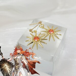 May include: A pair of gold-colored starburst earrings with a central orange gemstone. The earrings have a post-back closure and are displayed on a clear, angled surface. The starburst design features multiple pointed rays extending from the center.
