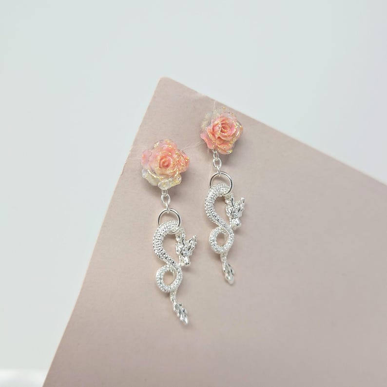 May include: A pair of dangle earrings featuring a pink and yellow rose stud, connected to a silver dragon charm. The dragon charm has a detailed, textured design with a dangling crystal at the bottom. The earrings are set against a soft pink and white background.