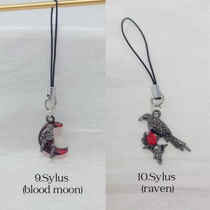 May include: Two phone charms with black cords and silver-tone metal hardware. The first charm, labeled 9.Sylus (blood moon), features a dark red crescent moon. The second charm, labeled 10.Sylus (raven), depicts a black raven perched on a branch with a red gem.