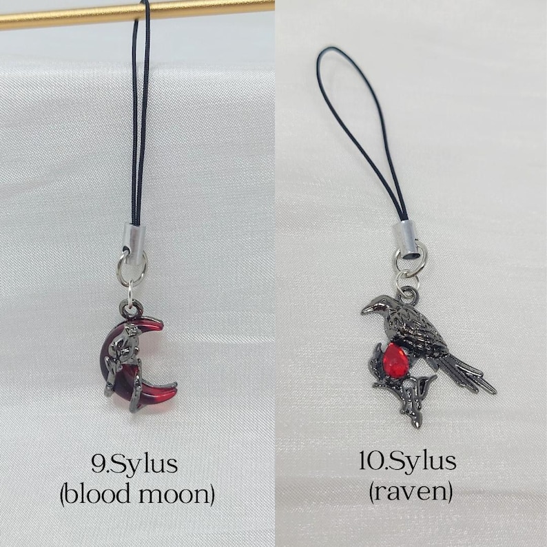 May include: Two phone charms with black cords and silver-tone metal hardware. The first charm, labeled 9.Sylus (blood moon), features a dark red crescent moon. The second charm, labeled 10.Sylus (raven), depicts a black raven perched on a branch with a red gem.