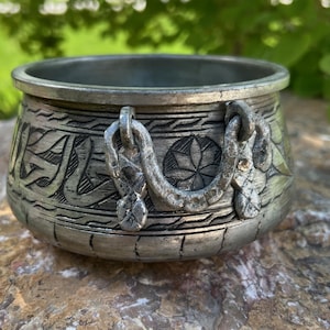 May include: A small, silver-colored metal bowl with intricate etched designs and two decorative chain handles. The bowl has a rounded shape and a slightly aged appearance, suggesting it may be vintage or antique. The background is blurred, with green foliage.