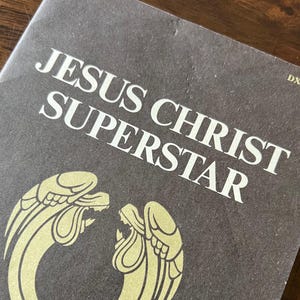 May include: A dark gray album cover with the title "JESUS CHRIST SUPERSTAR" in large white letters. Below the title is a gold illustration of two stylized angel wings. The album's code, DXSA-7206, is in the upper right corner.
